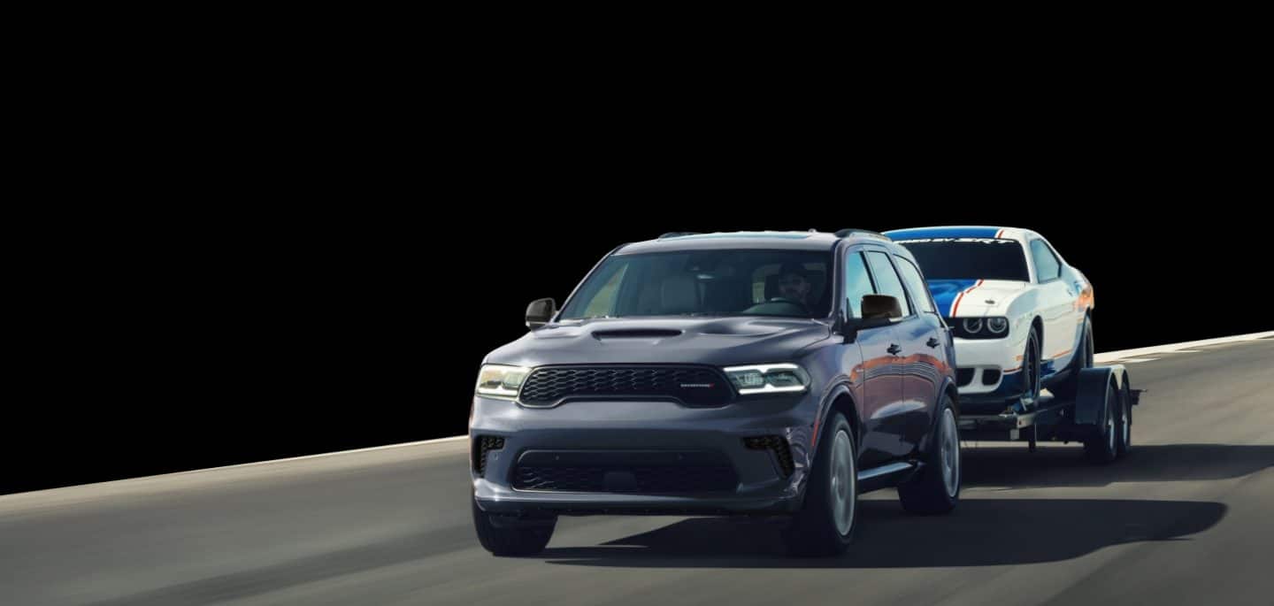 A gray 2026 Dodge Durango GT HEMI traveling down a road, towing a white Dodge Charger with a large blue racing center stripe. The background is blacked out.