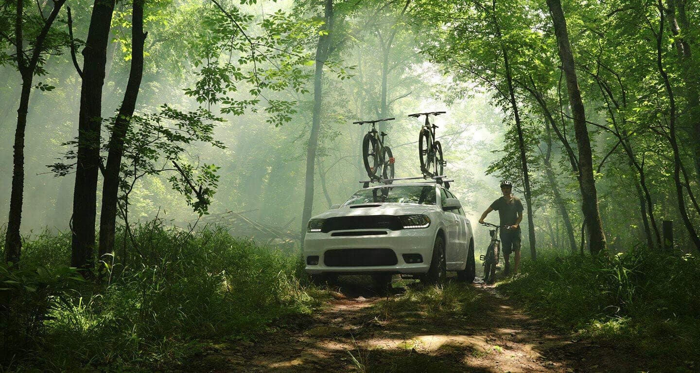 Display A three-quarter front view of a 2020 Dodge Durango, parked in the woods with two bicycles strapped to the roof rack.