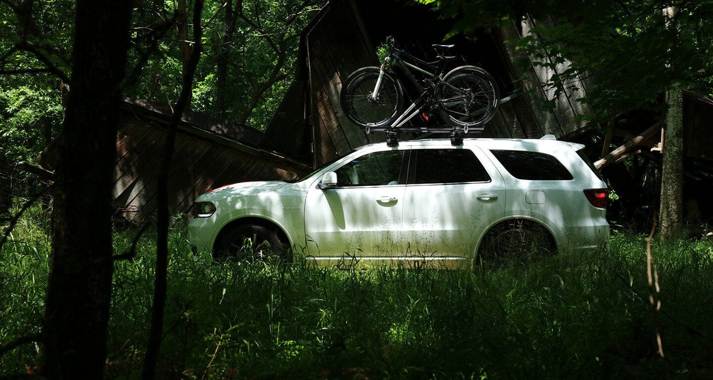 Display A profile view of a 2020 Dodge Durango parked in the woods with two bicycles strapped to the roof rack.