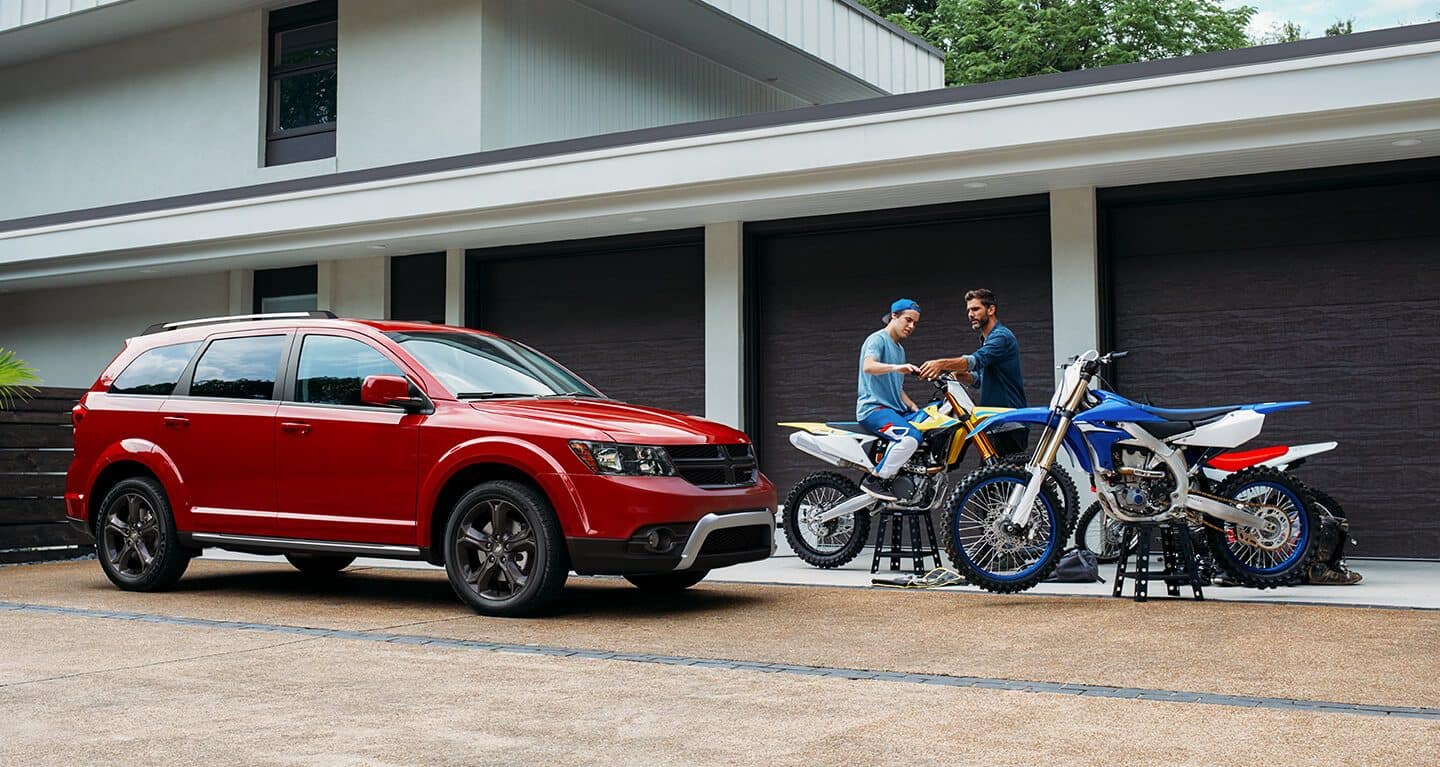 Display The 2020 Dodge Journey parked in a driveway beside two men working on dirtbikes raised on stands.