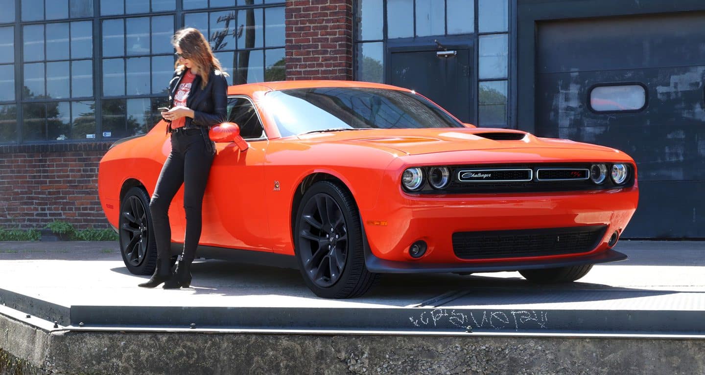 Display The 2021 Dodge Challenger R/T Scat Pack parked outside a warehouse with a woman leaning on its passenger door.