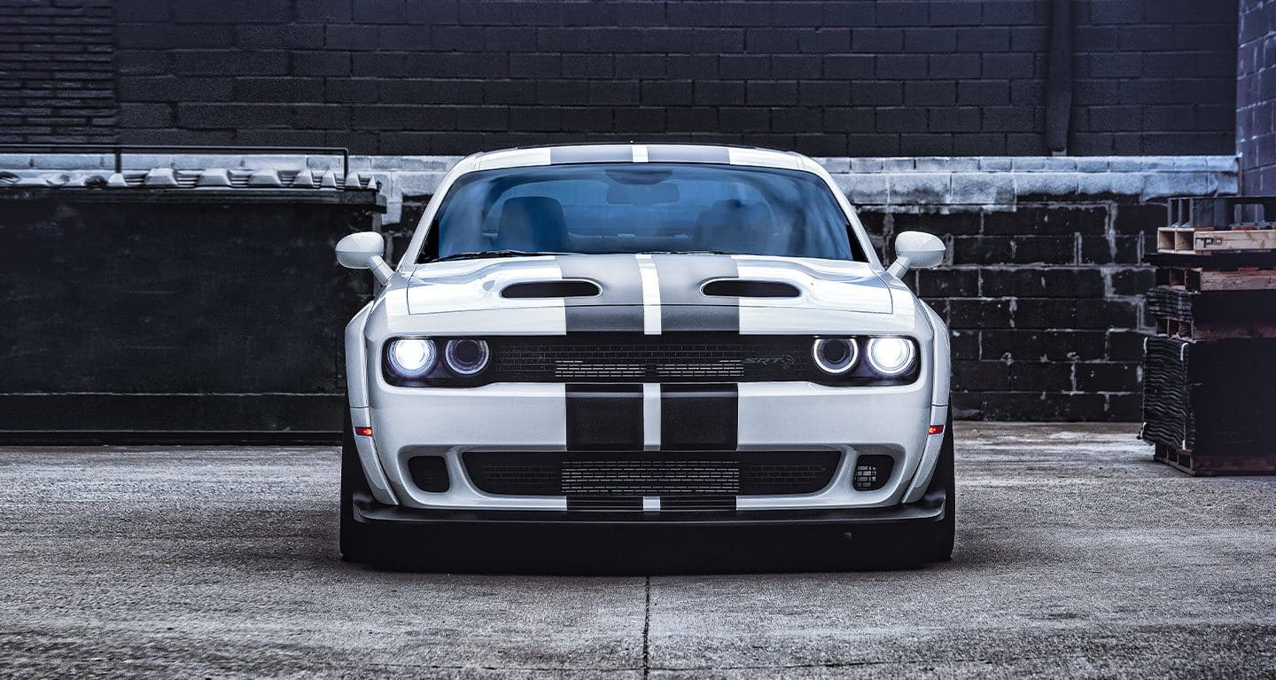 Display Front shot of a white 2022 Dodge Challenger SRT Hellcat Redeye Widebody in front of a black brick wall.