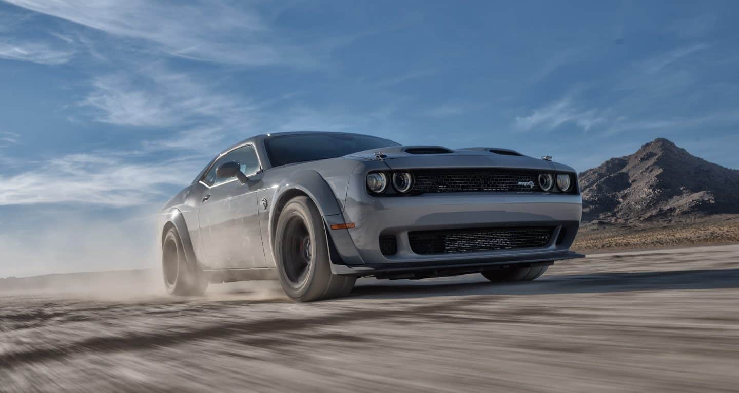 Display A gray 2022 Dodge Challenger SRT Super Stock driving on a dirt road with a mountain in the background.