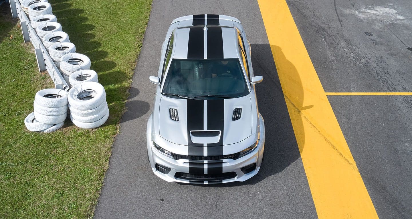 Display An overhead view of a 2022 Dodge Charger SRT Hellcat Widebody with twin center stripes, parked beside a fence of white painted tires.