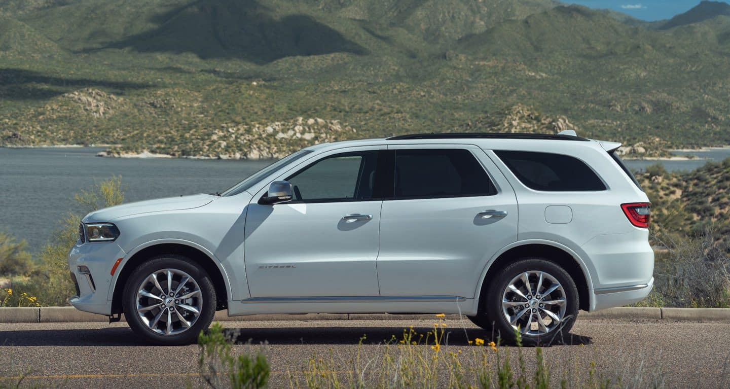 Display A passenger-side profile view of a white 2022 Dodge Durango Citadel parked beside a concrete wall, broken fence and ominous stormy sky.