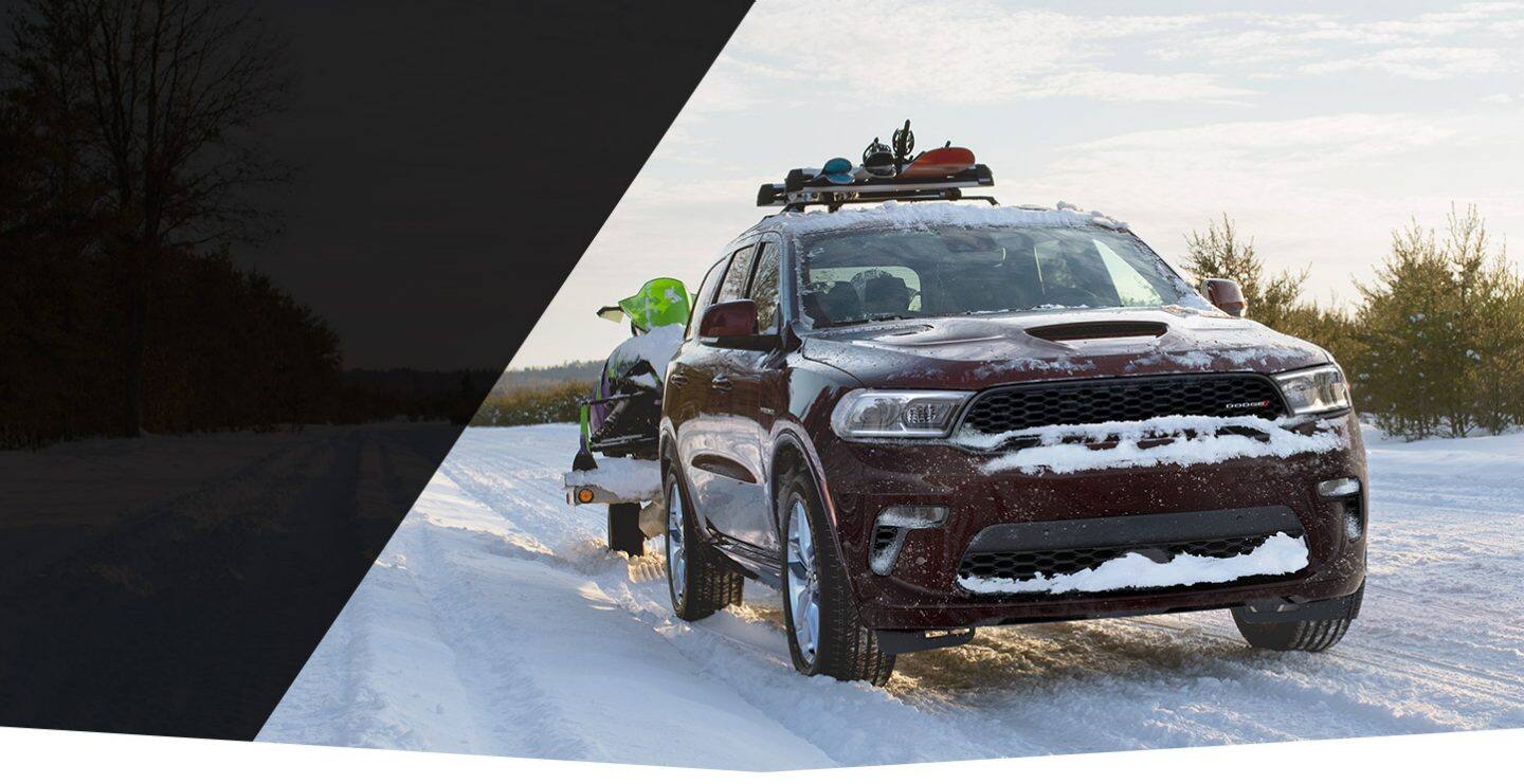 The 2022 Dodge Durango drives on a snowy trail with a snowmobile in tow and skis, poles and a snowboard on its roof rack.
