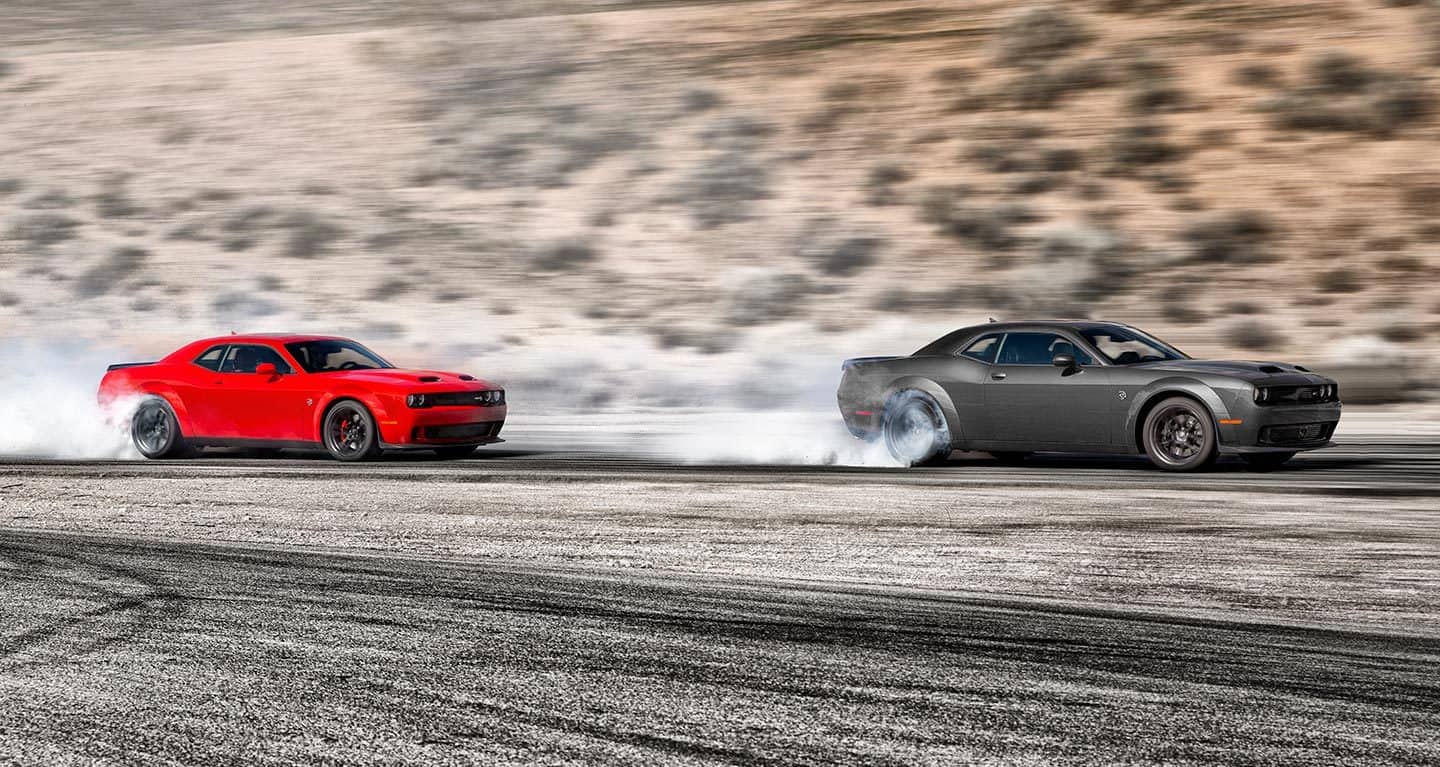 Display Two Dodge Challenger models on a track. In front, a gray 2023 Dodge Challenger SRT Hellcat, followed by a red 2023 Dodge Challenger SRT Hellcat, both with smoke emanating from their rear tires.