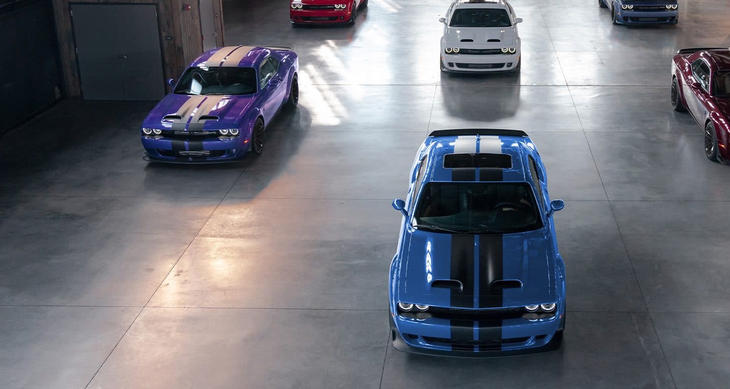 Display Six 2023 Dodge Challenger models parked in a commercial garage. They include a purple Challenger SRT Hellcat, a blue Challenger SRT Hellcat with black center stripes, a red Challenger SRT Hellcat, a white Challenger SRT Hellcat and more.