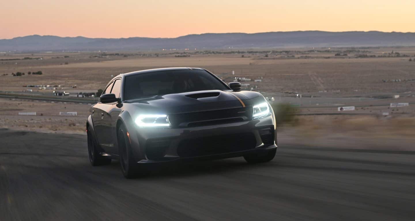 Display A 2023 Dodge Charger SRT Hellcat with its headlamps on, being driven on a track in the desert at dusk.