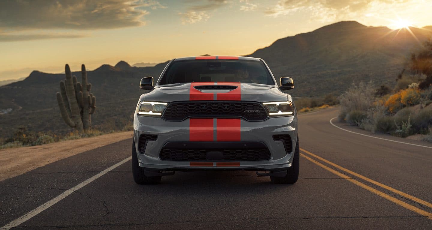 Display The 2023 Dodge Durango SRT 392 being driven on a desert road at sunset, its headlamps lit.