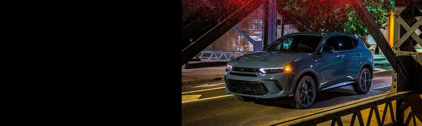 A 2023 Dodge Hornet R/T with the available Track Pack with its headlamps and cornering lamps on, being driven across a bridge at twilight.
