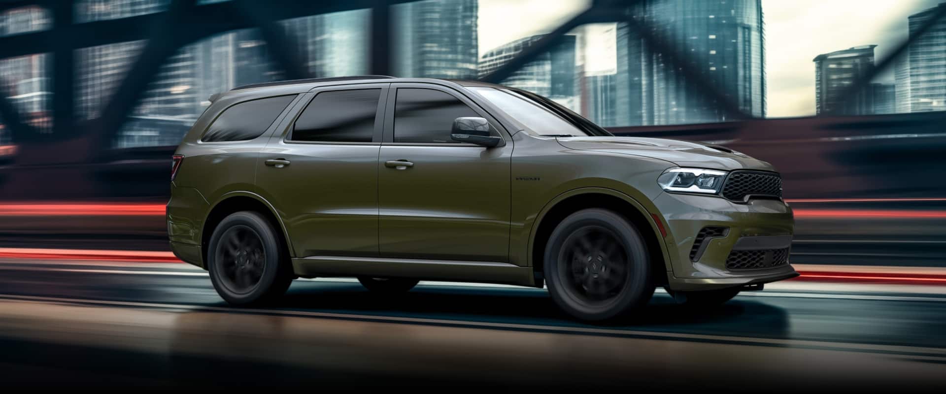 A passenger-side profile of a green 2026 Dodge Durango GT HEMI traveling across a bridge in a city with tall buildings in the background.