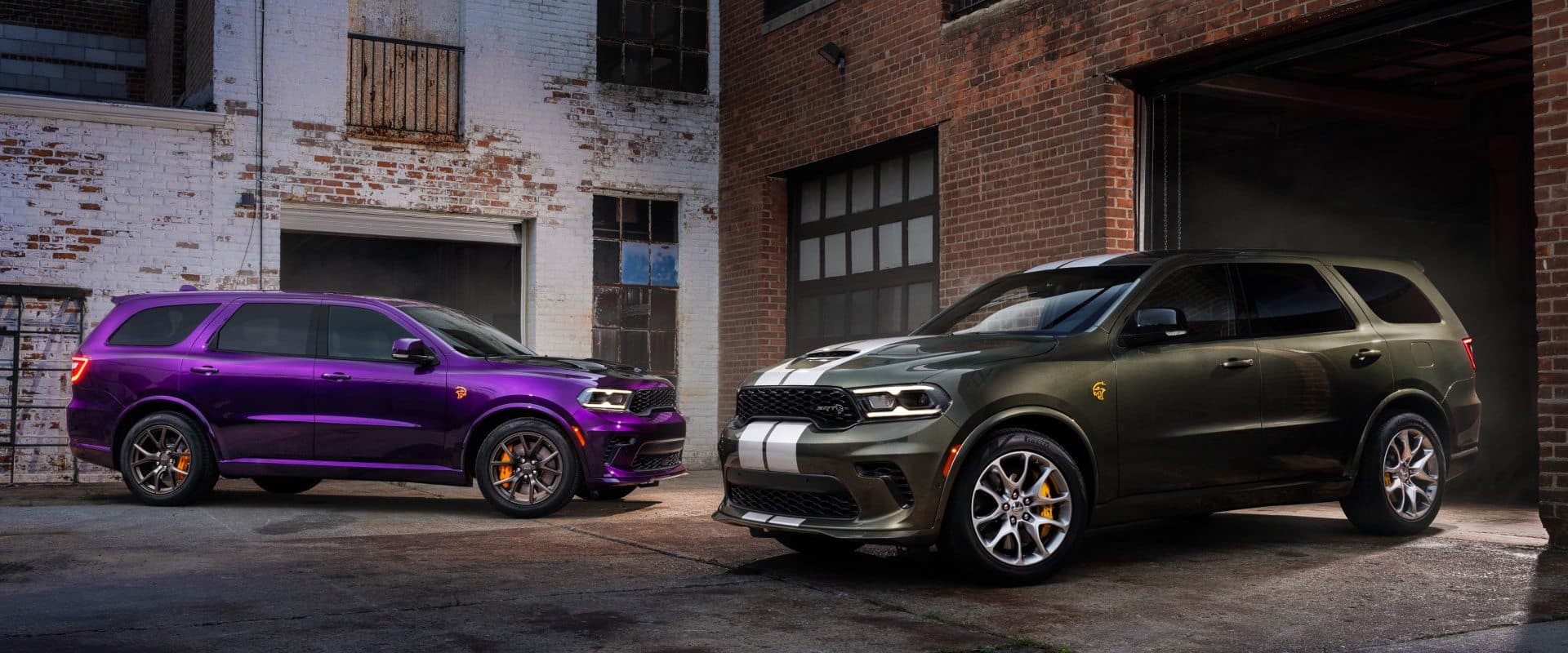 Two 2026 Dodge Durango models in an alley surrounded by brick buildings. On the left, a purple Durango SRT Hellcat Jailbreak with black painted hood is parked. On the right, a green Durango SRT Hellcat Jailbreak with silver dual center stripes is exiting a garage.