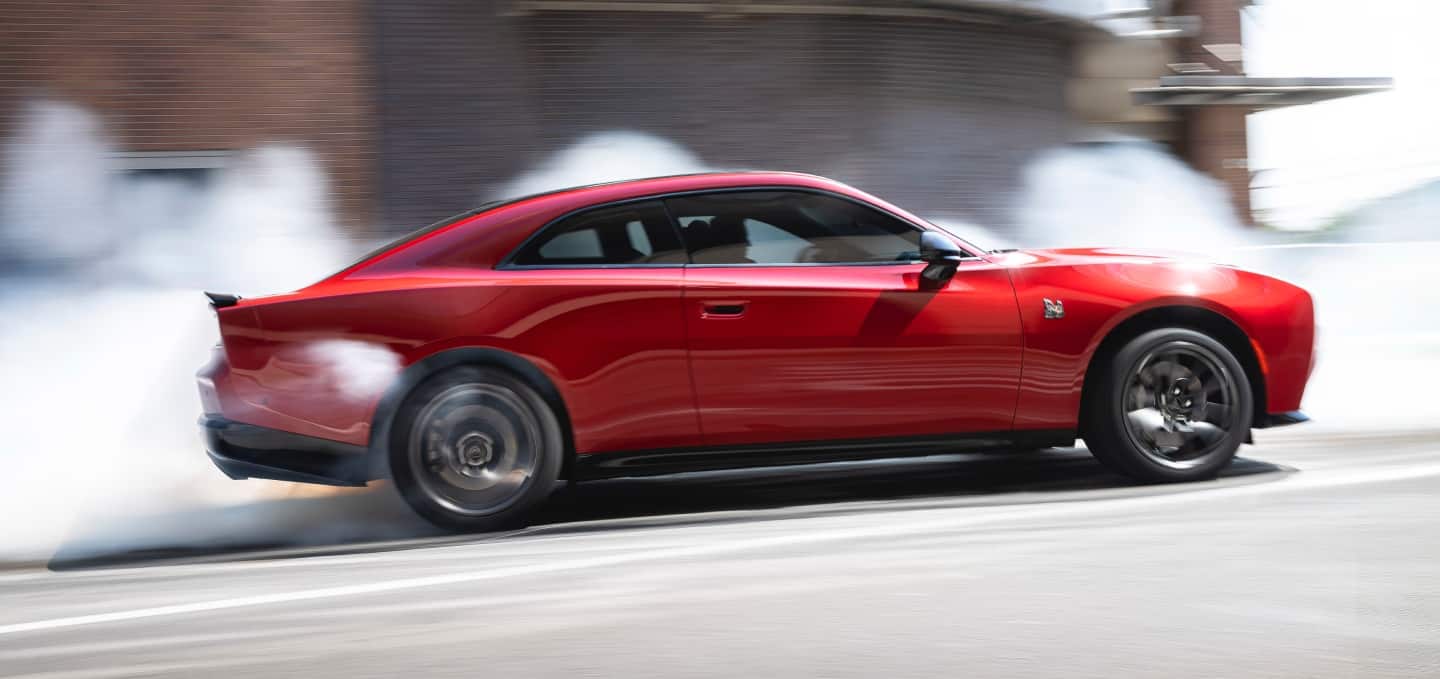 A red 2027 Dodge Charger Scat Pack in profile, stopped at an intersection, with smoke coming from its rear tires.