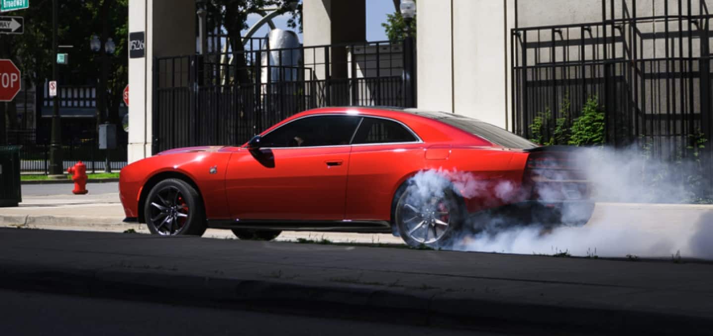 A red 2027 Dodge Charger Scat Pack in profile, stopped at an intersection, with smoke coming from its rear tires.