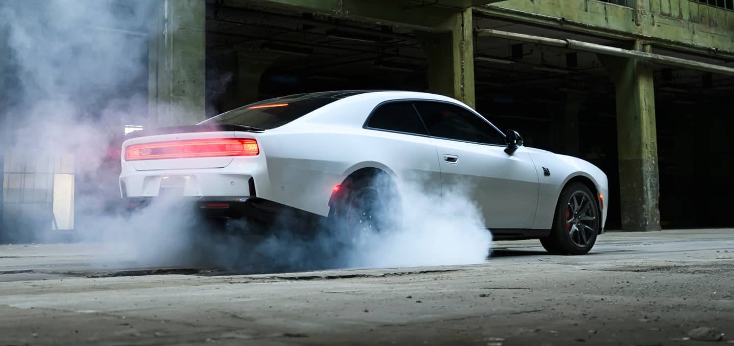 A white 2027 Dodge Charger Scat Pack, with smoke coming from its rear tires, shot from a rear profile.