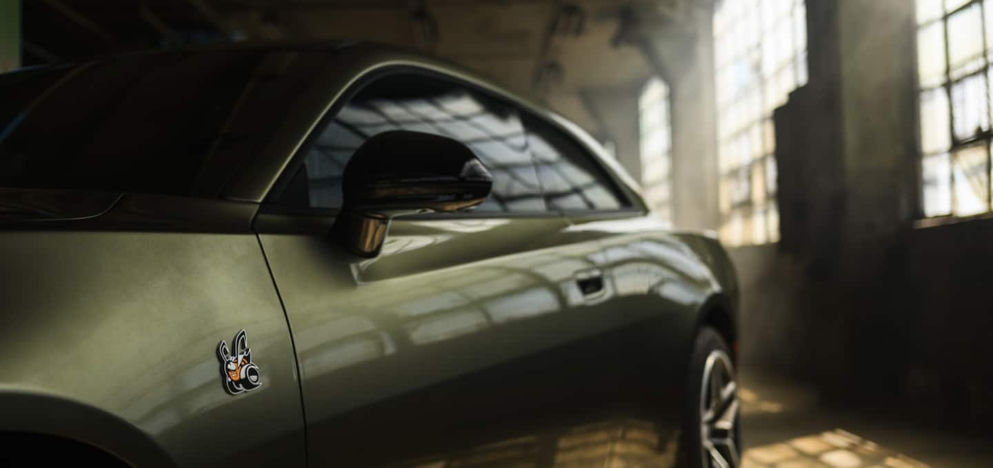 The driver's side of an olive green 2027 Dodge Charger Scat Pack, parked in an industrial garage.