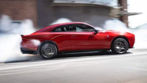 A red 2027 Dodge Charger Scat Pack in profile, stopped at an intersection, with smoke coming from its rear tires.