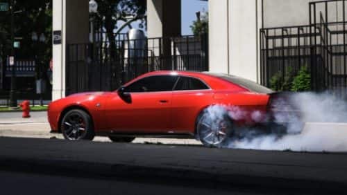 A red 2027 Dodge Charger Scat Pack in profile, stopped at an intersection, with smoke coming from its rear tires.