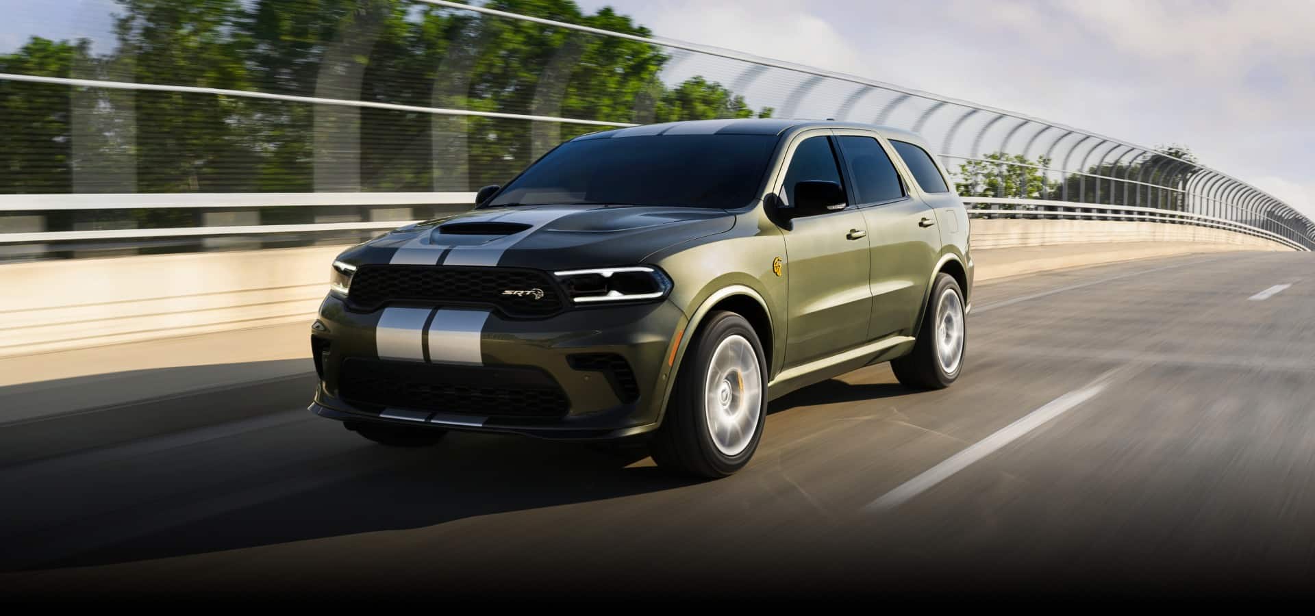 An olive green 2027 Dodge Durango SRT Hellcat Jailbreak with dual white center stripes, traveling over a highway bridge. Yes, It Rips.