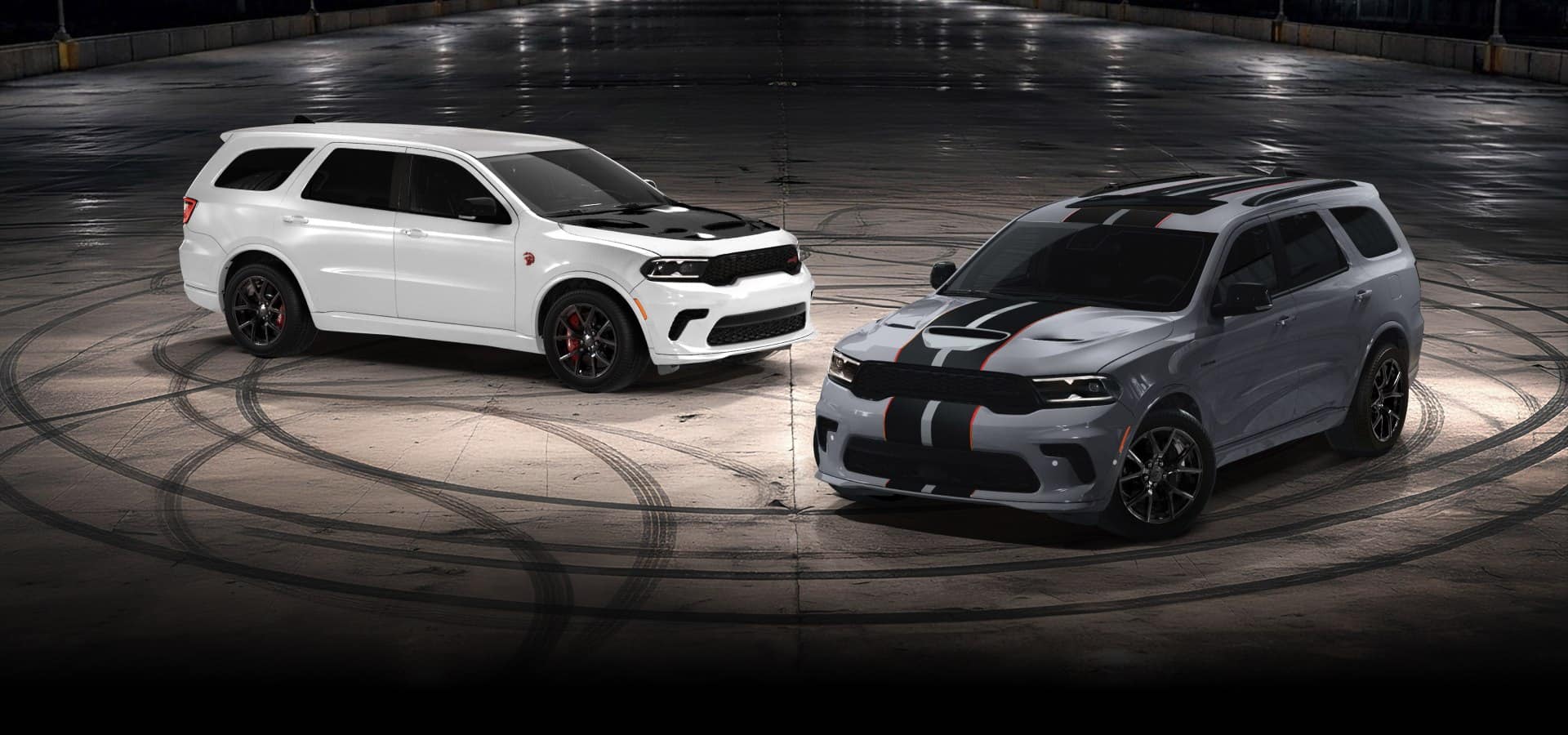 A white 2027 Dodge Durango SRT Hellcat Jailbreak with a black hood insert and a gray 2027 Dodge Durango GT Hemi V8 with dual black and red center stripes, parked perpendicular to each other on an empty lot at night. Smart and Seamless.