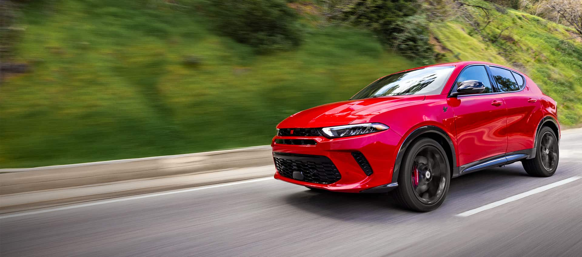 A front angled driver-side profile of a red 2024 Dodge Hornet RT being driven on a highway, with the background blurred to indicate the vehicle is in motion.