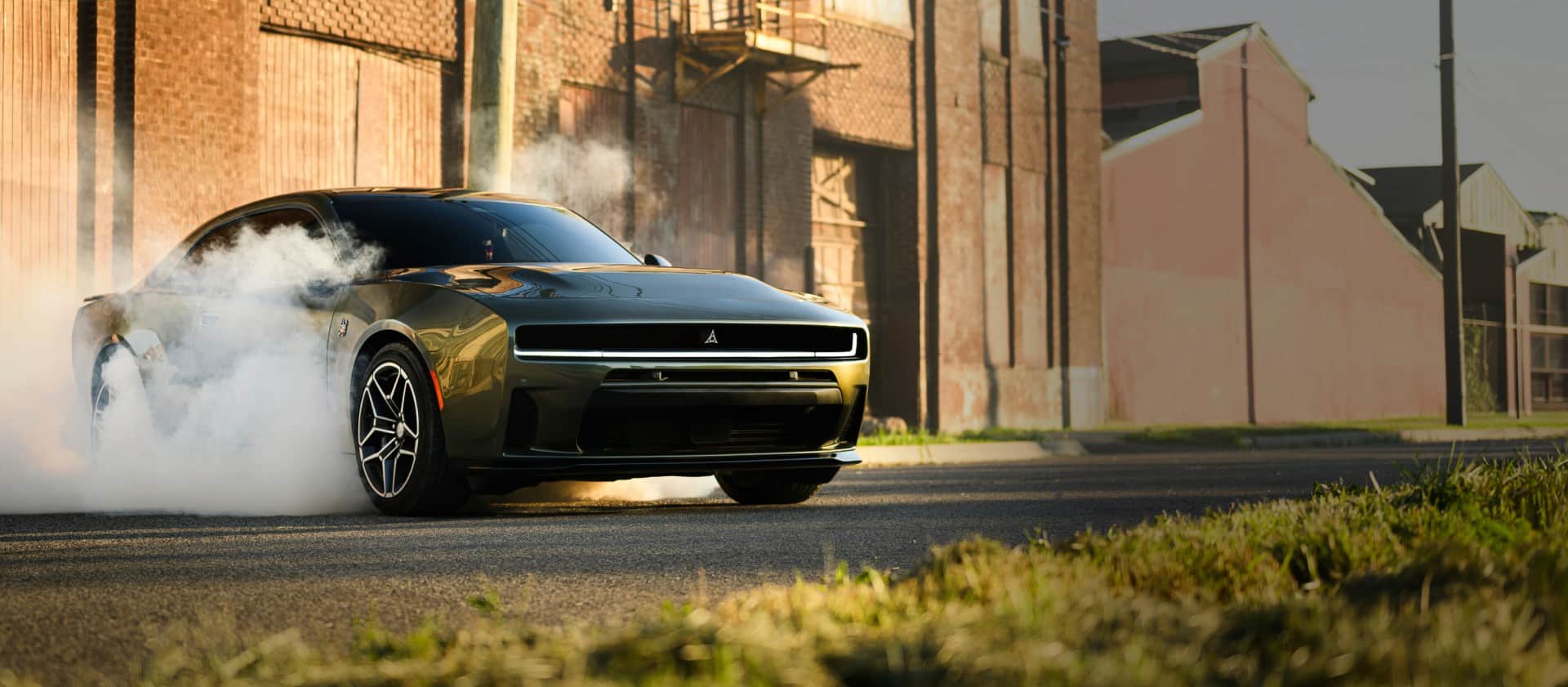 A passenger-side front angle of a black 2026 Dodge Charger Scat Pack Plus parked on blacktop with smoke billowing from its tires. In the background are industrial buildings.