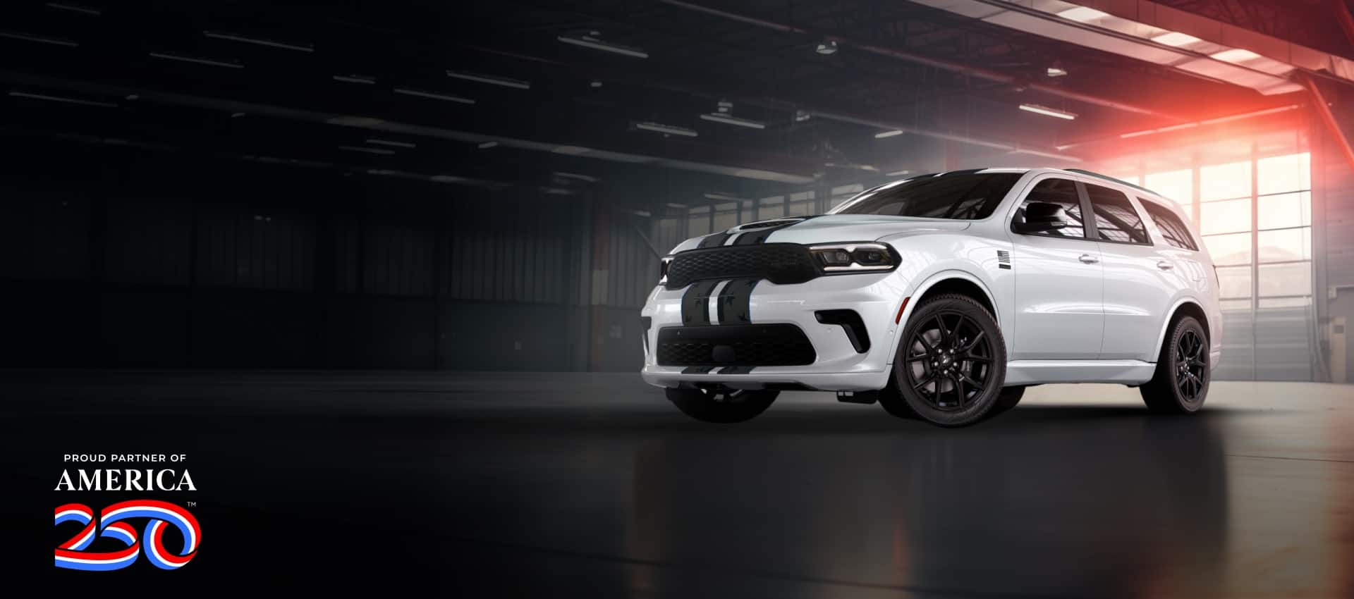 A white 2026 Dodge Durango GT America 250 with dual black center stripes, parked in a commercial garage. Proud Partner of America 250.