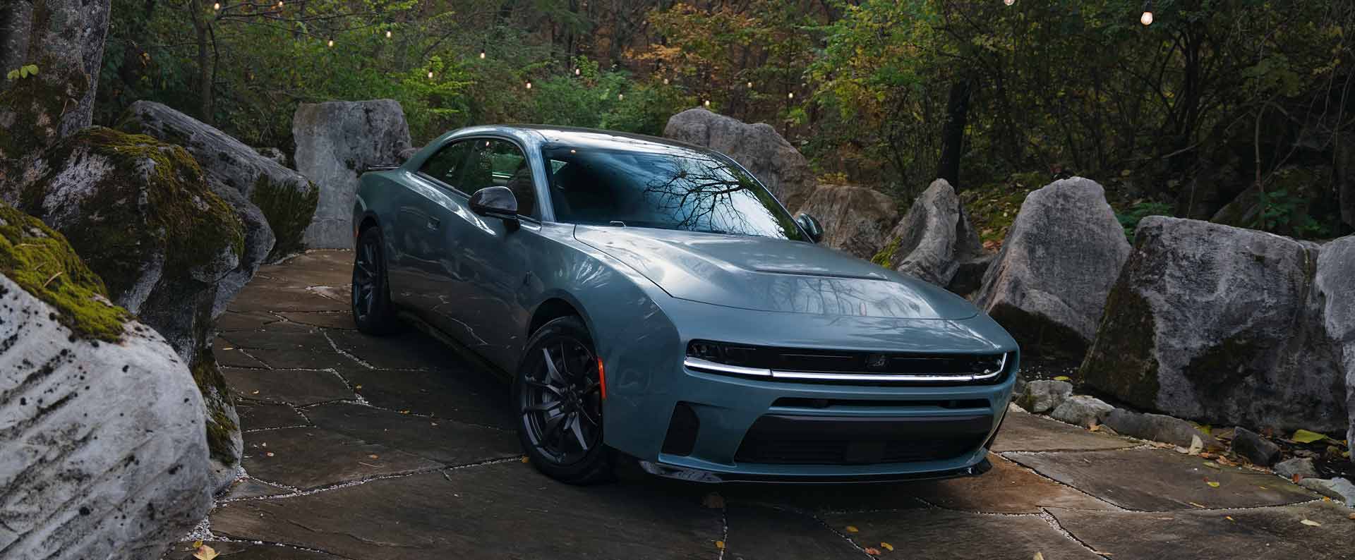 A two-door blue 2027 Dodge Charger Scat Pack on display surrounded by huge boulders and lush greenery.