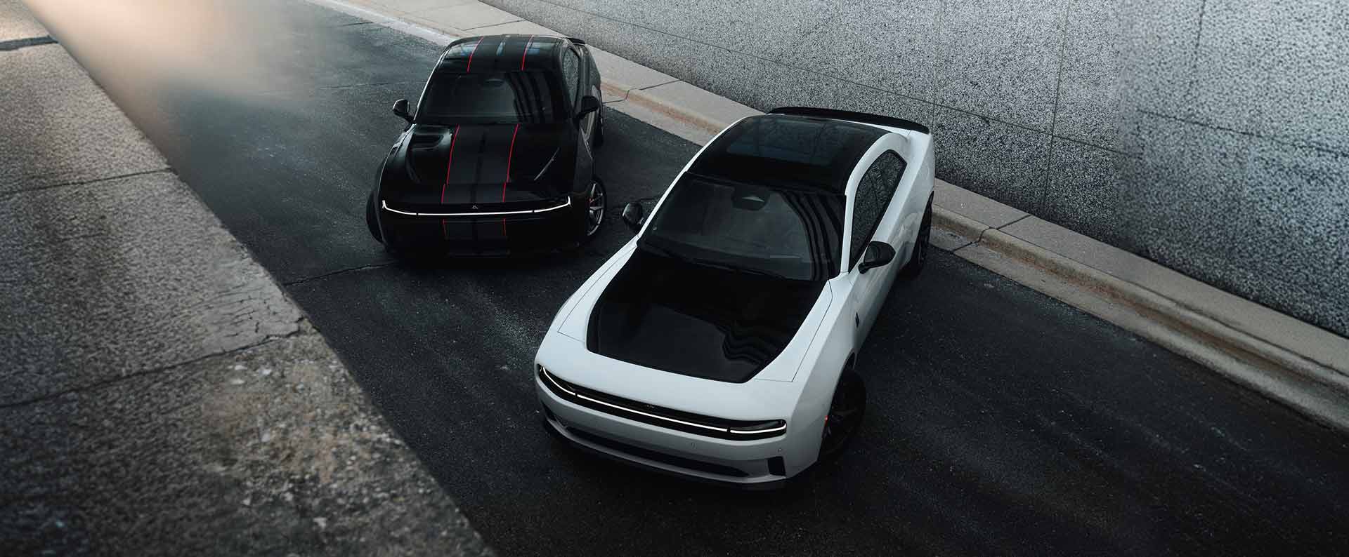 Two 2027 Dodge Charger two-door models: on the left, a black Charger Scat Pack with dual black and red center stripes and on the right, a white Charger Daytona Scat Pack with black hood and black roof, shot from above.