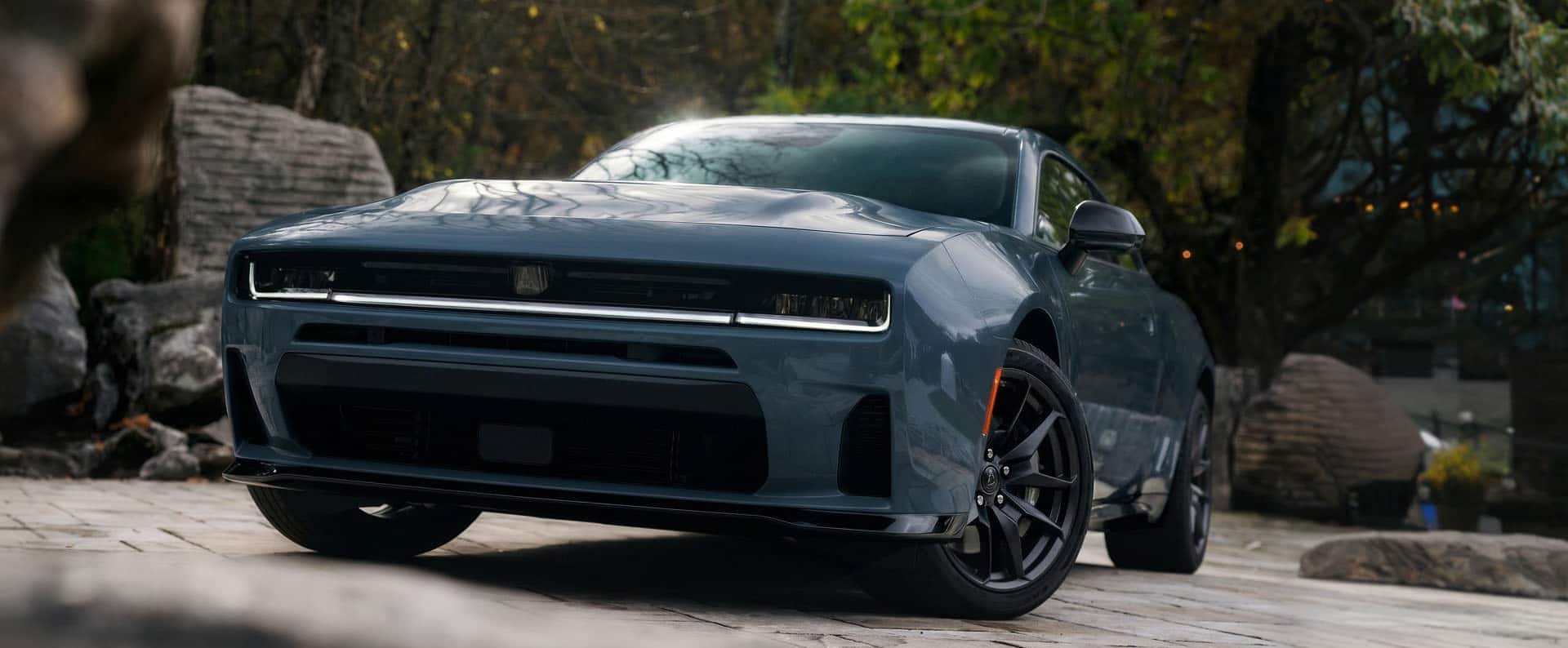 A two-door blue 2027 Dodge Charger Scat Pack on display surrounded by huge boulders and lush greenery.