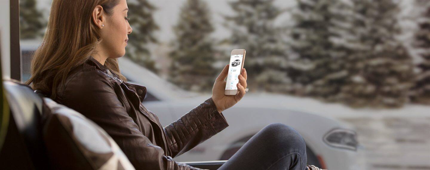 A woman sitting outdoors, looking at the Uconnect app on her smartphone, with her Dodge Charger in the background.