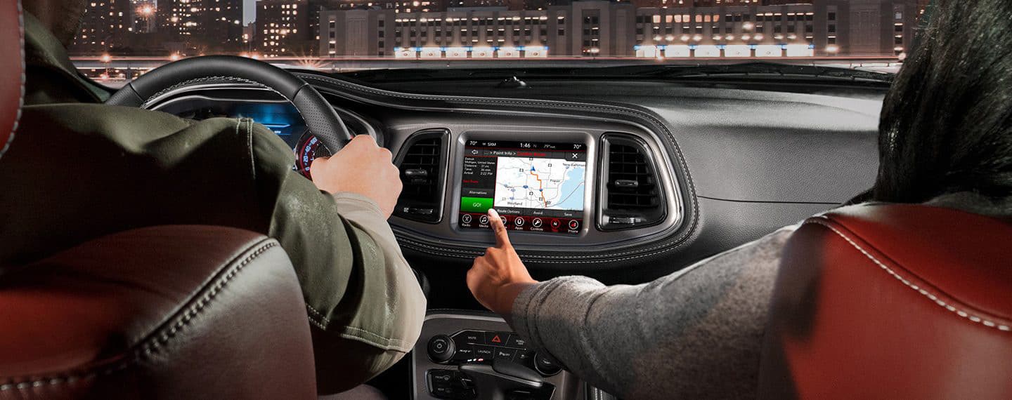 The front passenger in a 2021 Dodge Challenger reaching for the vehicle's touchscreen, on which a route map is displayed.