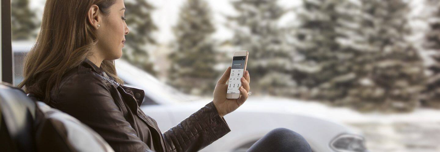 A woman sitting outdoors, looking at the Uconnect app on her smartphone, with her Dodge Charger in the background.
