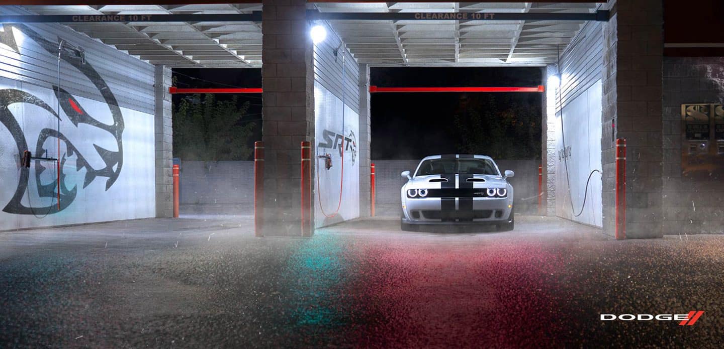 Display A silver 2021 Dodge Challenger SRT Hellcat Redeye Widebody with dual carbon center stripes parked, at night, in a self-serve car wash with SRT graphics on its walls.