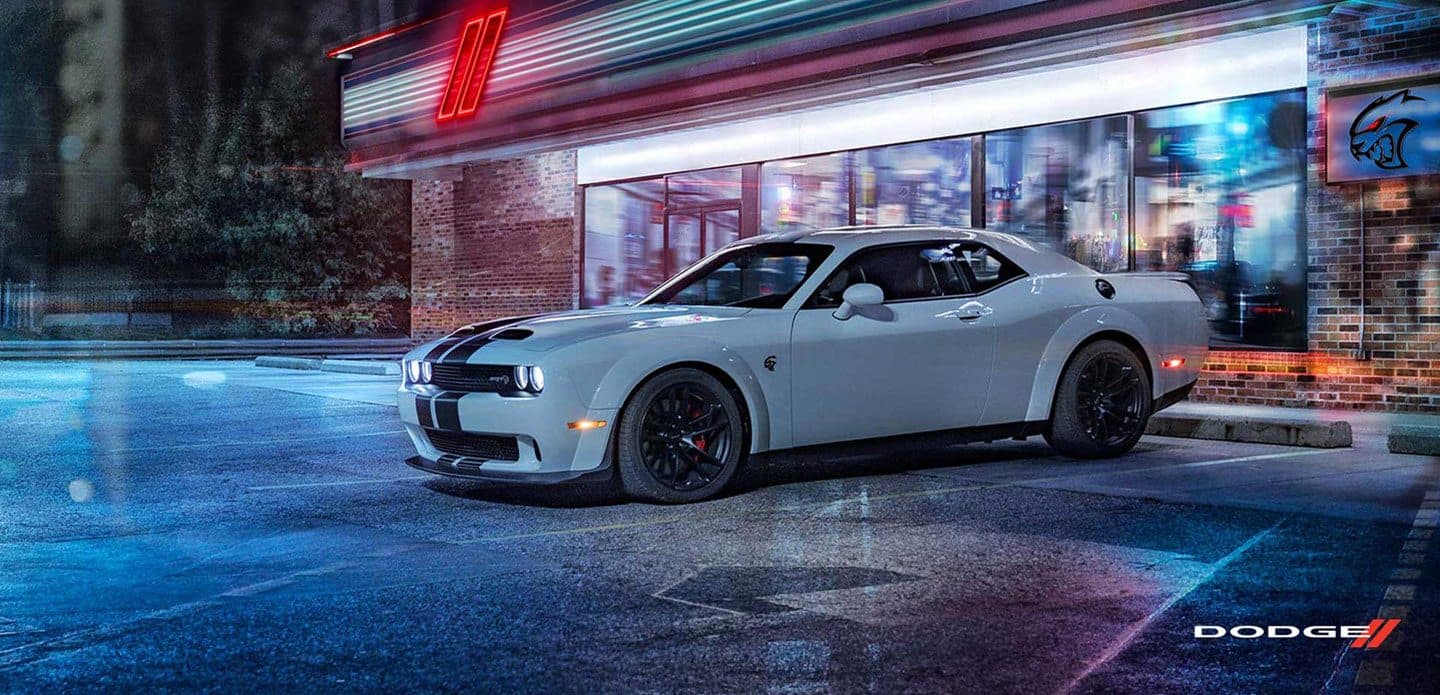 Display A silver 2021 Dodge Challenger SRT Hellcat Redeye Widebody with dual carbon center stripes parked, at night, in the lot of a small store with Dodge racing stripes on the store's neon sign.