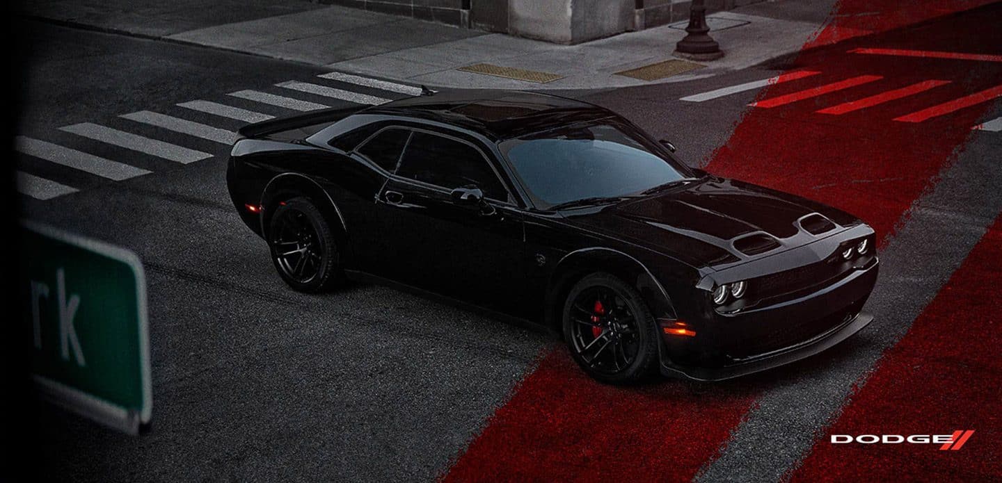 Display A black 2021 Dodge Challenger SRT Hellcat Widebody being driven through an intersection, at night, with the red Dodge racing stripes superimposed on the street.